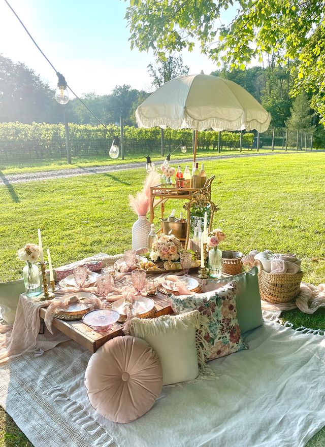 Boho-chic outdoor picnic in a sunlit vineyard: low wooden table on blankets with pastel plates and crystal glasses, floral cushions, candles, wicker basket, fringed umbrella and string lights.