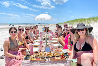 Sunny beach picnic brunch with a group of women in swimsuits seated around a long pink‑decorated table set with charcuterie boards, pastries, glassware and candles under a white parasol, with ocean waves and grassy sand dunes in the background.