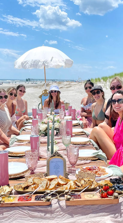 Group of smiling women in swimsuits gathered for a pink-themed beach picnic brunch around a low table with charcuterie boards, candles and a white parasol on a sunny sandy ocean beach with dunes and a pier in the distance.
