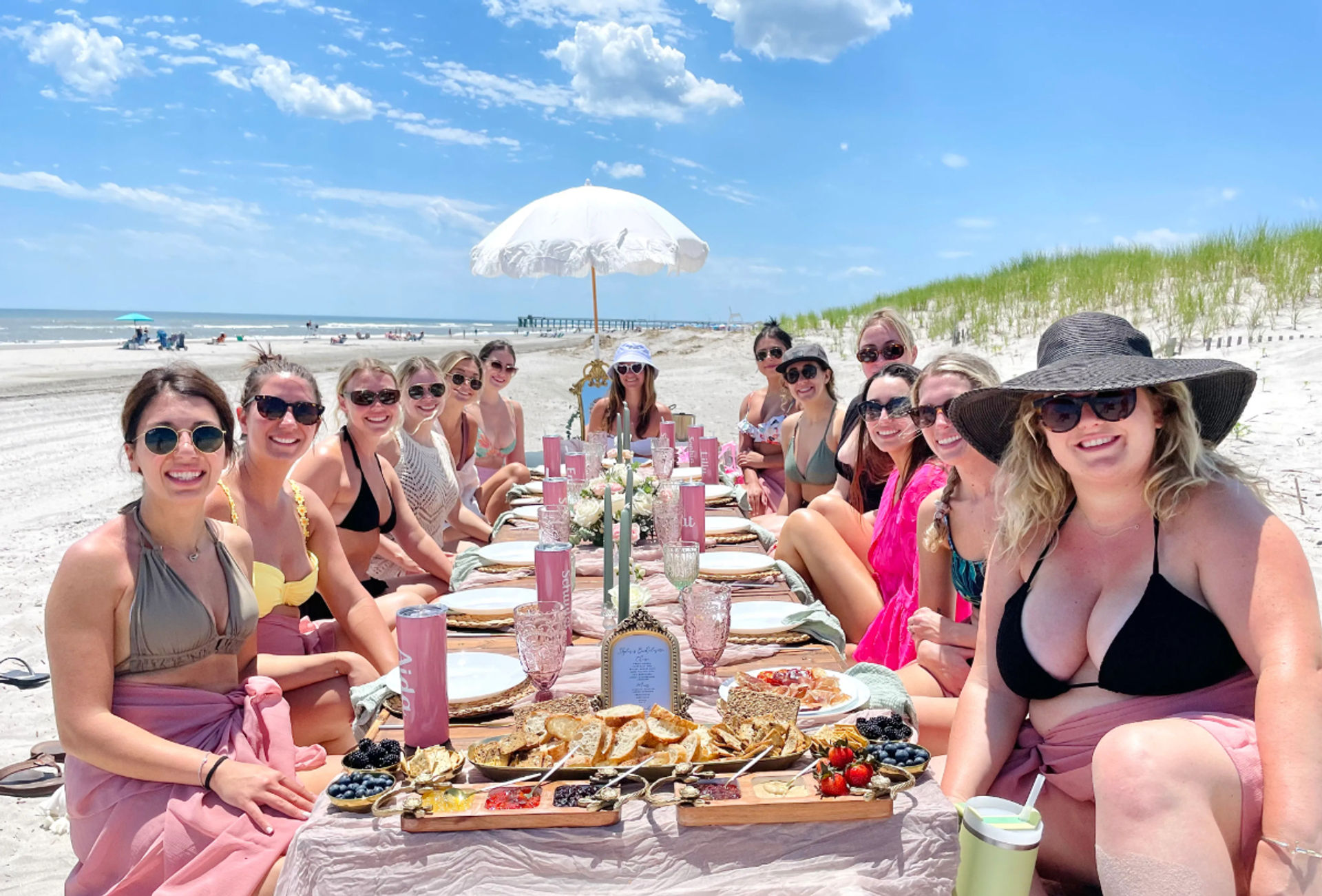 Group of smiling women in swimsuits gathered for a pink-themed beach picnic brunch around a low table with charcuterie boards, candles and a white parasol on a sunny sandy ocean beach with dunes and a pier in the distance.