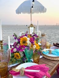 Sunset beach picnic with a boho low wooden table, white fringed umbrella, vibrant sunflower bouquet, colorful glass goblets, taper candles, woven placemats and bright cloth napkins.