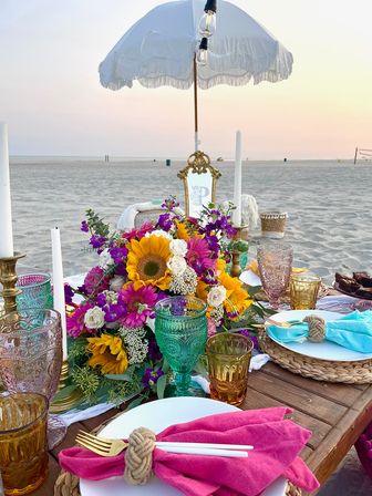 Sunset beach picnic with a boho low wooden table, white fringed umbrella, vibrant sunflower bouquet, colorful glass goblets, taper candles, woven placemats and bright cloth napkins.
