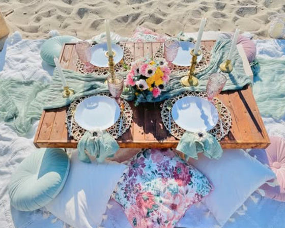 Boho beach picnic on sand: low wooden table set with white plates on woven chargers, gold candlesticks, pink goblets, pastel linens and floral cushions around a colorful flower centerpiece.