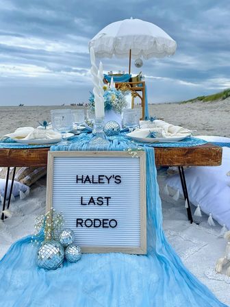 Beach picnic scene on sand with a low wooden table under a white parasol, blue linens and plates, crystal glassware, twisted white candles, floral centerpiece, small mirrored disco balls, and a letter board reading "LAST RODEO".