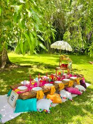 Boho-style backyard picnic on a sunny green lawn under a shady tree: low wooden table with colorful pom-pom pillows, bright napkins, candles, glassware, floral centerpiece and white parasol.