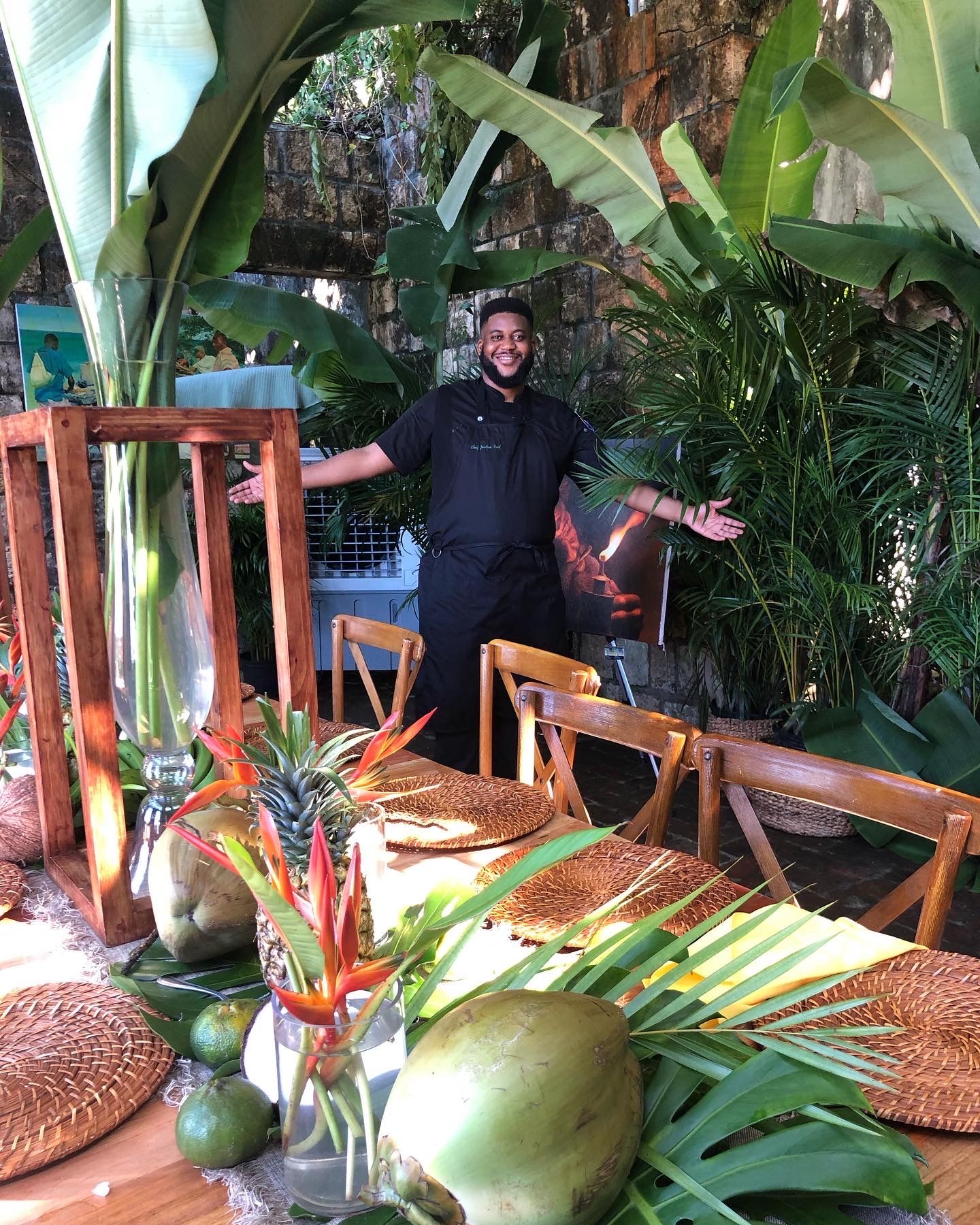 Smiling chef with arms open behind a tropical outdoor dining table on a rustic stone patio, table styled with coconuts, pineapple, banana leaves, bird-of-paradise flowers and woven placemats amid lush greenery.
