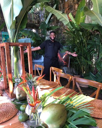 Smiling chef with arms open behind a tropical outdoor dining table on a rustic stone patio, table styled with coconuts, pineapple, banana leaves, bird-of-paradise flowers and woven placemats amid lush greenery.