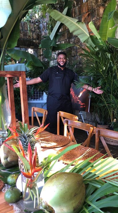 Smiling chef with arms open behind a tropical outdoor dining table on a rustic stone patio, table styled with coconuts, pineapple, banana leaves, bird-of-paradise flowers and woven placemats amid lush greenery.