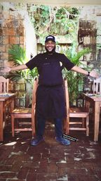 Smiling chef in black apron and cap stands with arms open between wooden tables on a brick patio in a lush, rustic outdoor courtyard with stone walls and tropical plants.