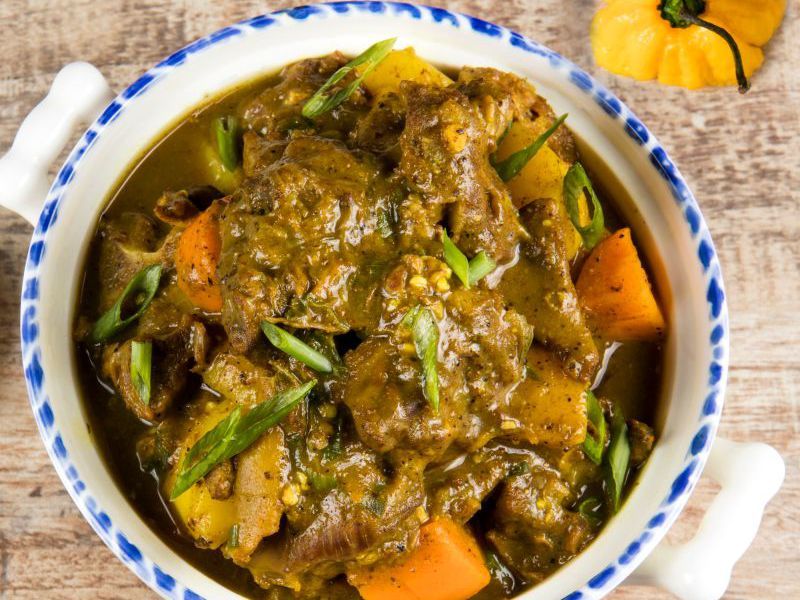 Hearty braised meat stew with chunky potatoes and carrots in a rich brown gravy, garnished with sliced green onions in a blue‑rimmed bowl on a rustic wooden table