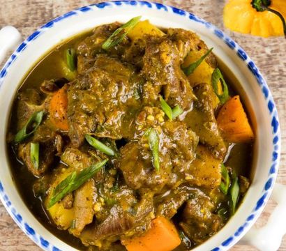Hearty braised meat stew with chunky potatoes and carrots in a rich brown gravy, garnished with sliced green onions in a blue‑rimmed bowl on a rustic wooden table
