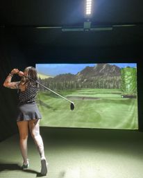Tattooed woman golfer in follow-through swinging a driver inside an indoor golf simulator, big screen showing a mountain fairway and green turf mat