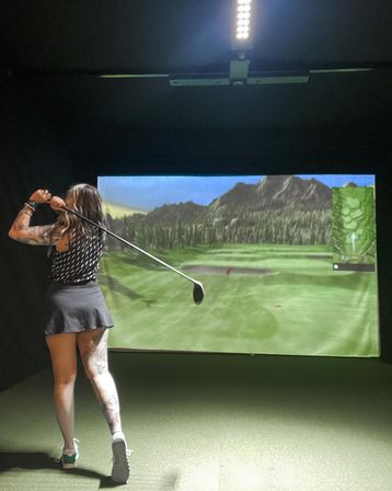 Tattooed woman golfer in follow-through swinging a driver inside an indoor golf simulator, big screen showing a mountain fairway and green turf mat