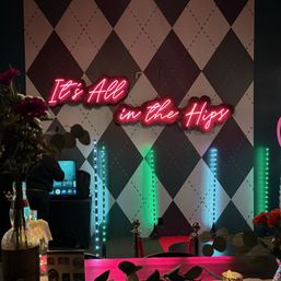 Pink neon sign reading 'It's All in the Hips' on a black-and-white diamond wall above a lit photo-booth style event backdrop with vertical LED tubes, red carpet and stanchions, flowers in the foreground