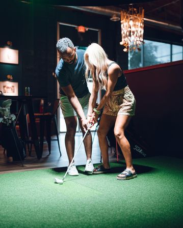 Hands-on putting lesson on an indoor artificial turf green — instructor guiding a woman’s grip at a casual urban indoor golf facility with clubs nearby.