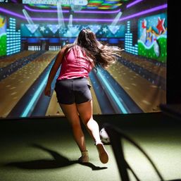 Person bowling in an indoor virtual bowling alley, releasing a ball down colorful neon-lit lanes toward an animated star mural