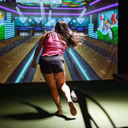 Person bowling in an indoor virtual bowling alley, releasing a ball down colorful neon-lit lanes toward an animated star mural
