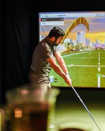 Golfer taking a swing in an indoor golf simulator on green turf, virtual driving range with city skyline and a yellow arch on the screen