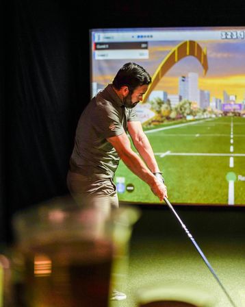 Golfer taking a swing in an indoor golf simulator on green turf, virtual driving range with city skyline and a yellow arch on the screen
