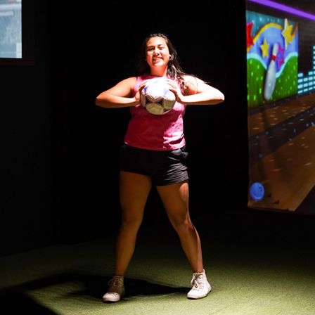 Smiling young woman holding a soccer ball on indoor turf under a spotlight, colorful arcade-style mural in the background.