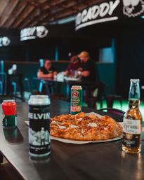 Cheesy pizza on a paper-lined plate with assorted beer cans and a bottled lager on a dark table in a cozy indoor pub, blurred patrons chatting in the background