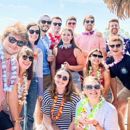 Group of young adults wearing colorful leis and sunglasses at a sunny waterfront summer party, smiling and holding drinks under a thatched umbrella