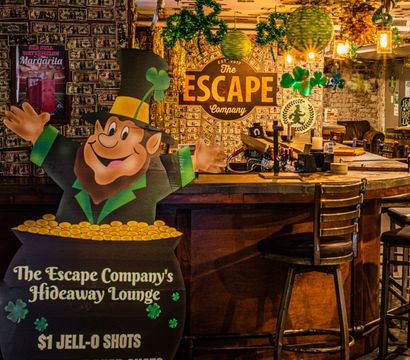 Cozy Irish-themed bar lounge with a cartoon leprechaun and pot of gold, shamrock decorations, dollar-bill–covered walls and wooden bar stools.
