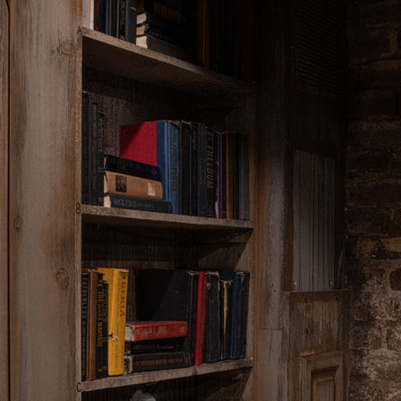 Moody close-up of a rustic wooden bookshelf filled with worn, colorful hardcover books beside an exposed brick wall — cozy vintage reading nook.