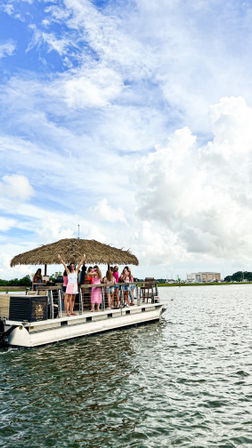 Pontoon boat with a thatched tiki roof carrying a group of friends celebrating near a coastal marina — sunny blue sky, fluffy clouds, and rippling water.