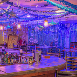 Ocean-themed bar interior with curved wooden bar, hanging inflatable sharks, blue fairy lights, bubble decorations, and wooden tables and chairs