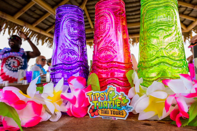 Bright purple, pink, and green translucent tiki cups on a wooden bar, surrounded by pink-and-white plumeria leis and a playful tiki-tour sticker beneath a thatched tiki-hut roof.