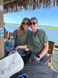 Two friends in matching green bachelorette shirts and sunglasses smiling under a tiki thatch roof on a sunny waterfront dock, with coastal marsh and blue sky behind them and purse, phone and drinks on the table.