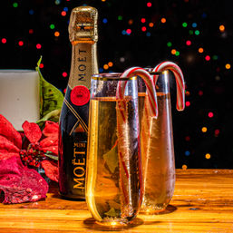 Festive Christmas table: two sparkling wine flutes garnished with candy canes, a mini bottle, red poinsettia and candle on a wooden table against colorful bokeh lights