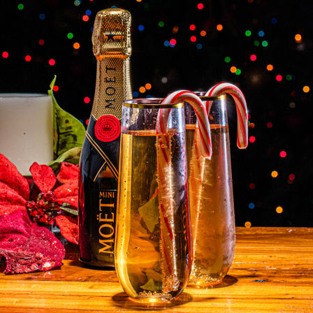 Festive Christmas table: two sparkling wine flutes garnished with candy canes, a mini bottle, red poinsettia and candle on a wooden table against colorful bokeh lights