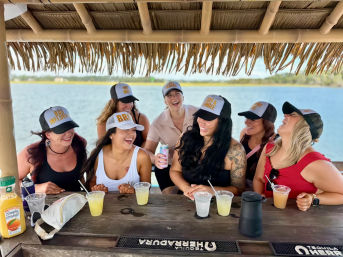 Seven friends laughing under a thatched tiki hut at a sunny waterfront bar, wearing novelty trucker hats and sipping cocktails over a wooden counter
