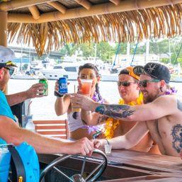 Friends toasting with cocktails and a canned beer under a thatched tiki-roof at a marina, yachts in the background for a summer waterfront party vibe.