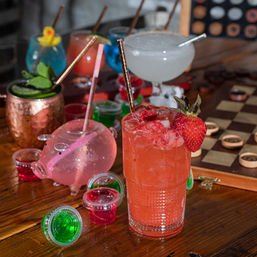 Playful lineup of craft cocktails at a bar: a bright strawberry highball with crushed ice and fresh strawberry, surrounded by colorful jello shots, a pink pig-shaped drink vessel, a copper Moscow mule with mint, and a frosty margarita by a wooden checkerboard.