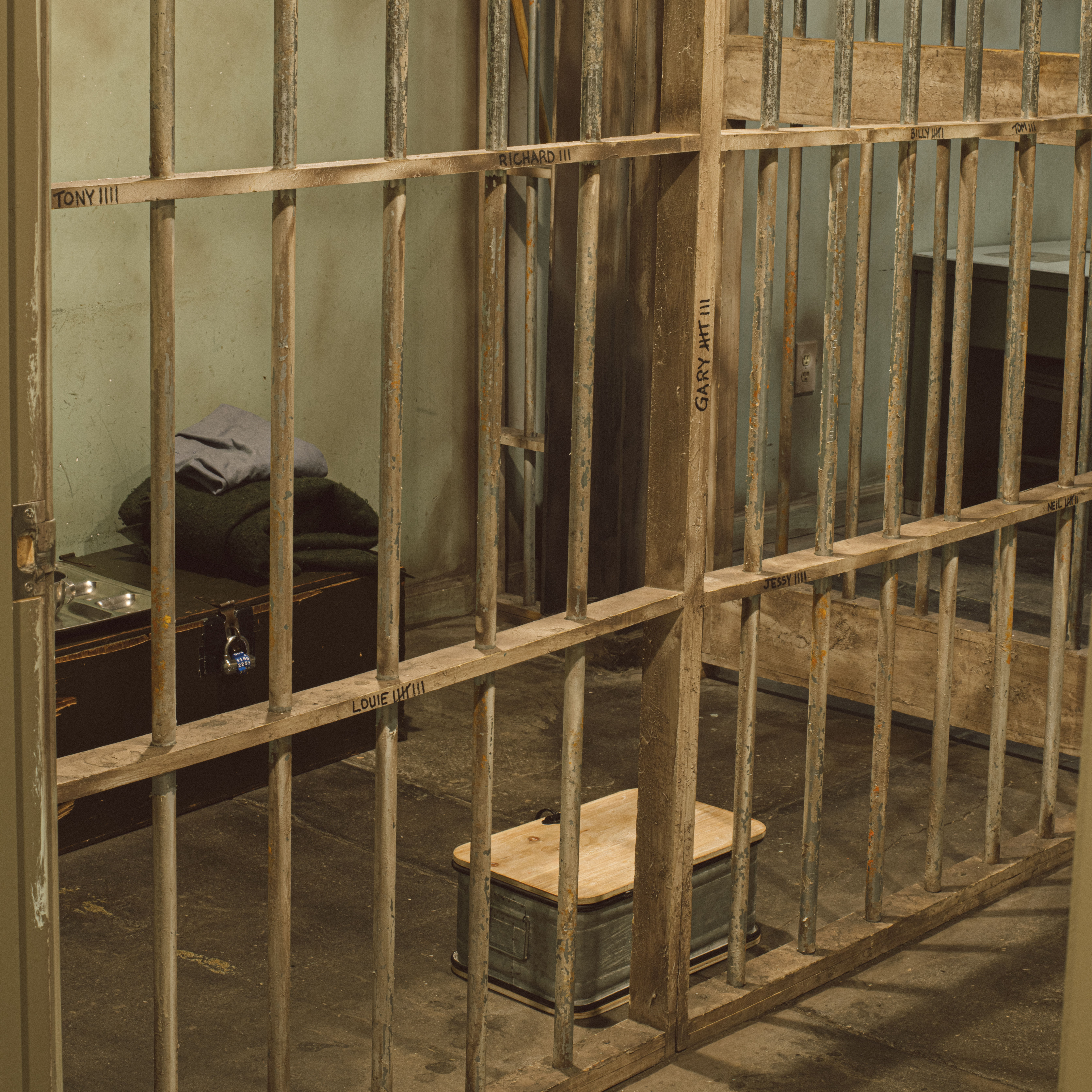 Weathered jail cell interior with rusted metal bars, cot with folded blanket, metal wash basin and small wooden stool