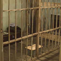 Weathered jail cell interior with rusted metal bars, cot with folded blanket, metal wash basin and small wooden stool