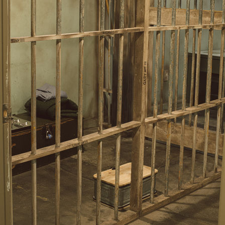 Weathered jail cell interior with rusted metal bars, cot with folded blanket, metal wash basin and small wooden stool