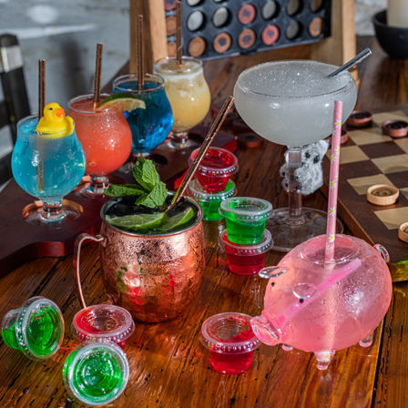 Colorful game-night cocktail spread on a wooden table at a cozy bar: blue, red and yellow stemmed cocktails (one with a rubber duck), a frosty margarita, a copper mug with mint and lime, stacked red and green jello shots, and a pink pig-shaped drink cup beside a checkers board.