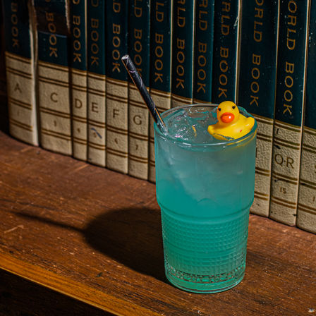 Turquoise iced cocktail in a textured glass with a metal straw and a tiny yellow rubber duck garnish, sitting on a wooden shelf in front of vintage encyclopedia spines.