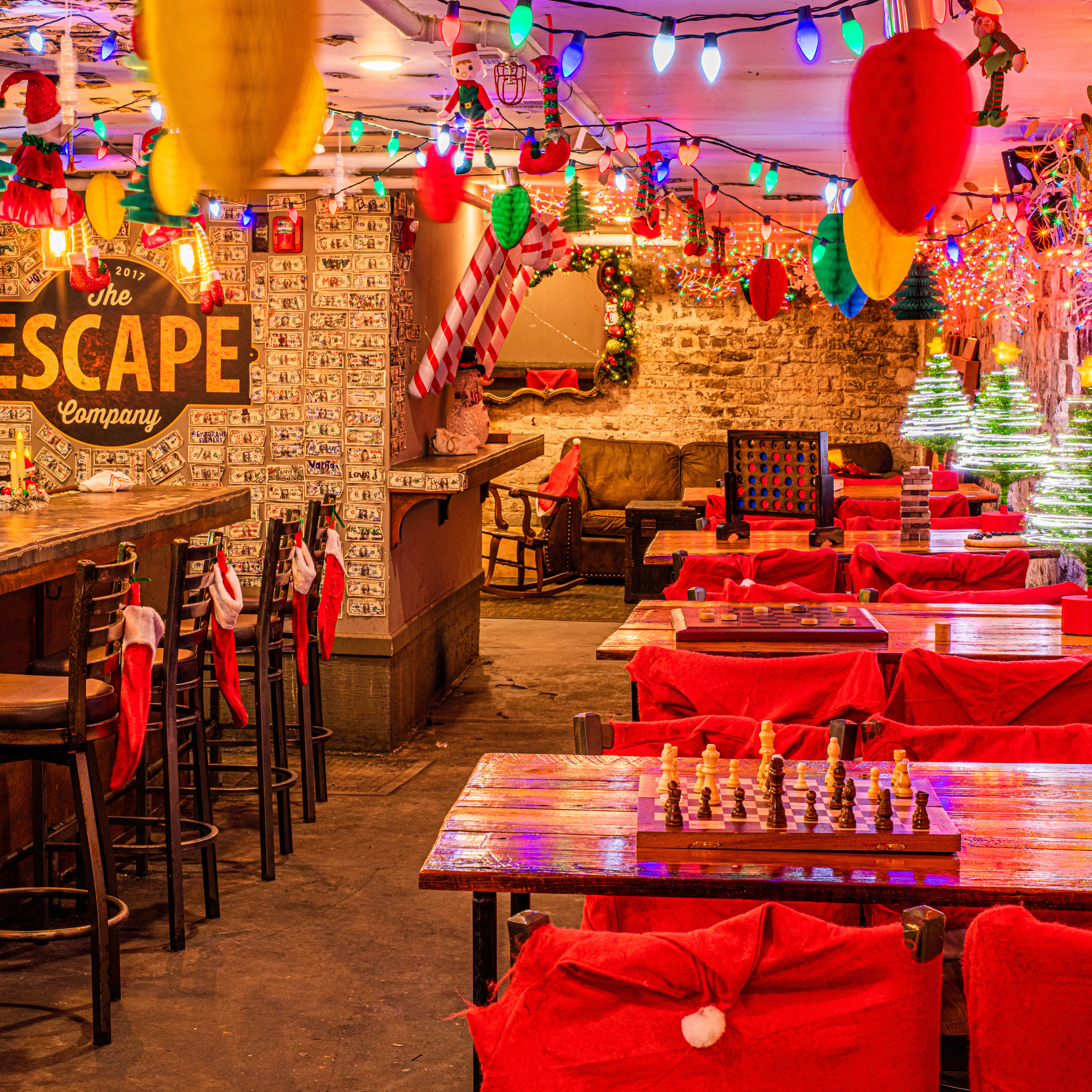 Cozy holiday-themed basement bar with colorful string lights, red chair covers and stockings, wooden tables set with chess and board games, candy-cane accents and lit mini Christmas trees.
