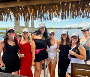 Laughing group of seven women at a waterfront tiki hut bachelorette party — bride in a white swimsuit and friends in matching caps holding small cups, bridge and marsh in the background