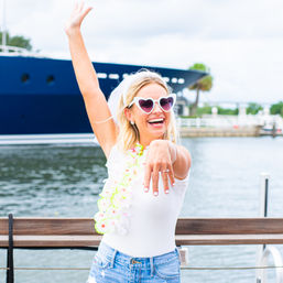 Cheerful woman at a marina wearing heart-shaped sunglasses, a white veil and floral lei, proudly showing an engagement ring with a yacht and water in the background.