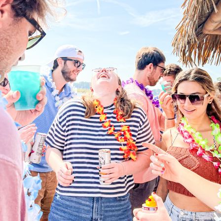 Sunny tropical-themed beach party with young adults wearing colorful leis and sunglasses, laughing and holding canned drinks under a tiki umbrella
