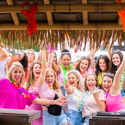 Energetic group of women in bright summer outfits and flower leis laughing, raising hands, and posing under a thatched tiki hut at a tropical-themed outdoor summer party.