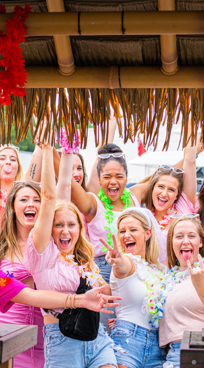 Energetic group of women in bright summer outfits and flower leis laughing, raising hands, and posing under a thatched tiki hut at a tropical-themed outdoor summer party.