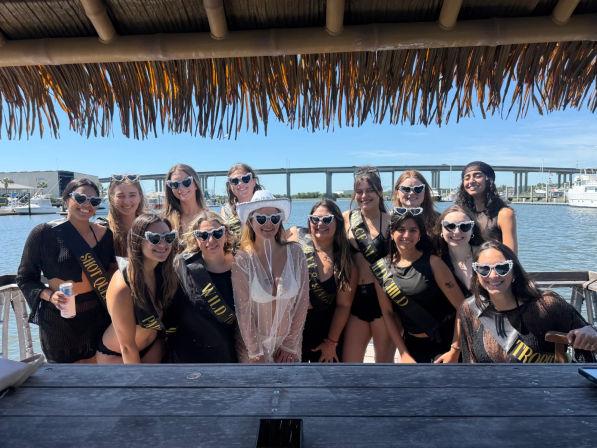 Group of women posing under a tiki thatched roof at a sunny marina, wearing matching heart-shaped sunglasses and black sashes for a bachelorette celebration, bride in a white hat and sheer cover-up, boats and a bridge visible over the water in the background