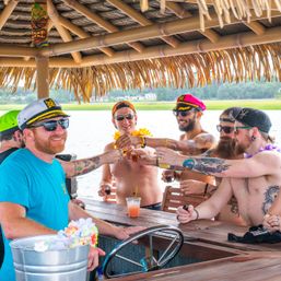 Group of friends in captain hats and sunglasses, shirtless with tattoos, toasting cocktails under a tiki-roofed floating bar on a sunny waterfront marsh — boat party vibes.